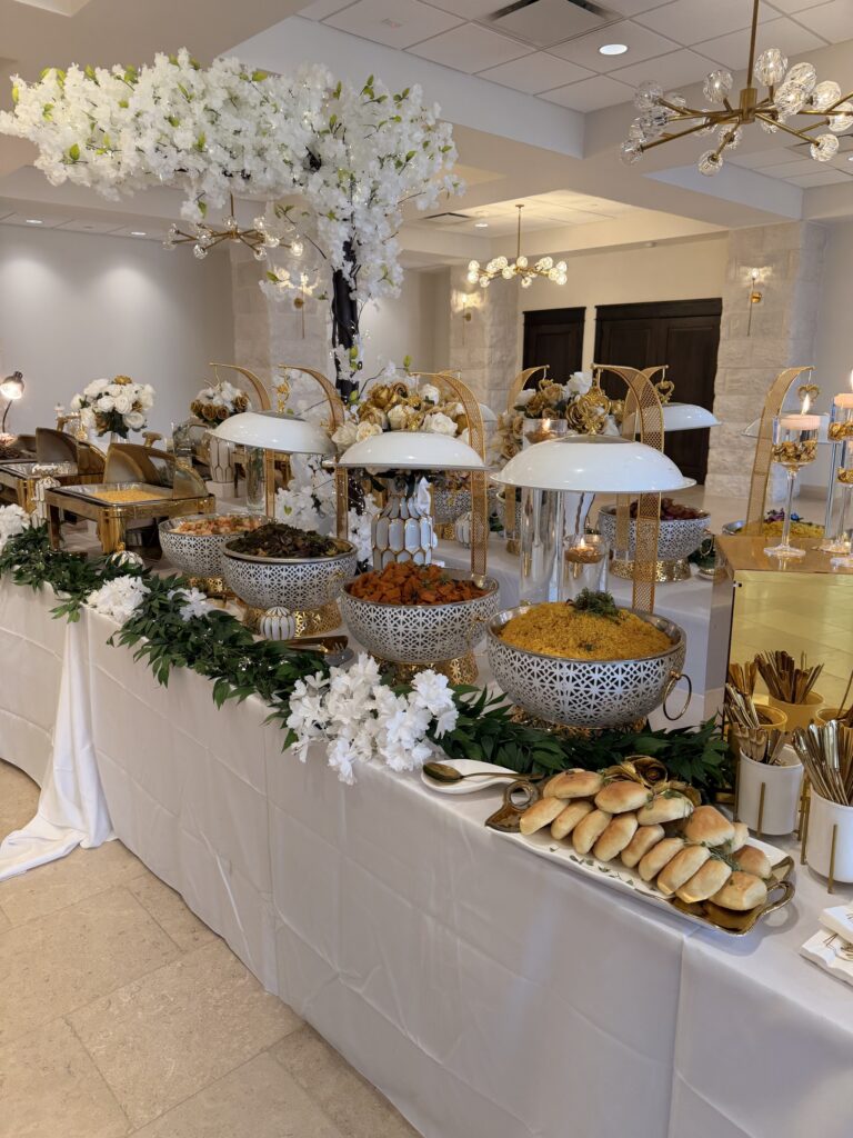 An indoor catering buffet with open patterned serving bowls, yellow rice, bread rolls, gold accents, white flowers and greenery.