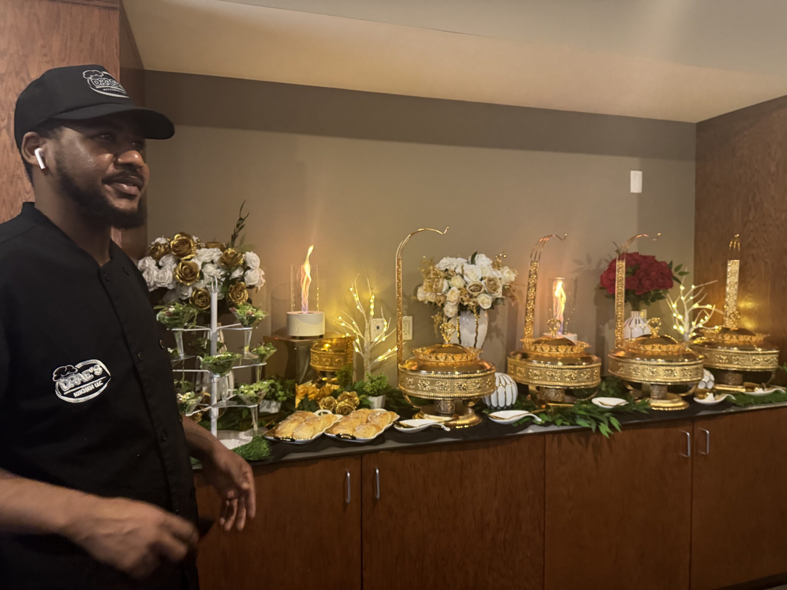 Chef Deandrae beside a gold buffet display decorated with flowers, greenery and tall candles.