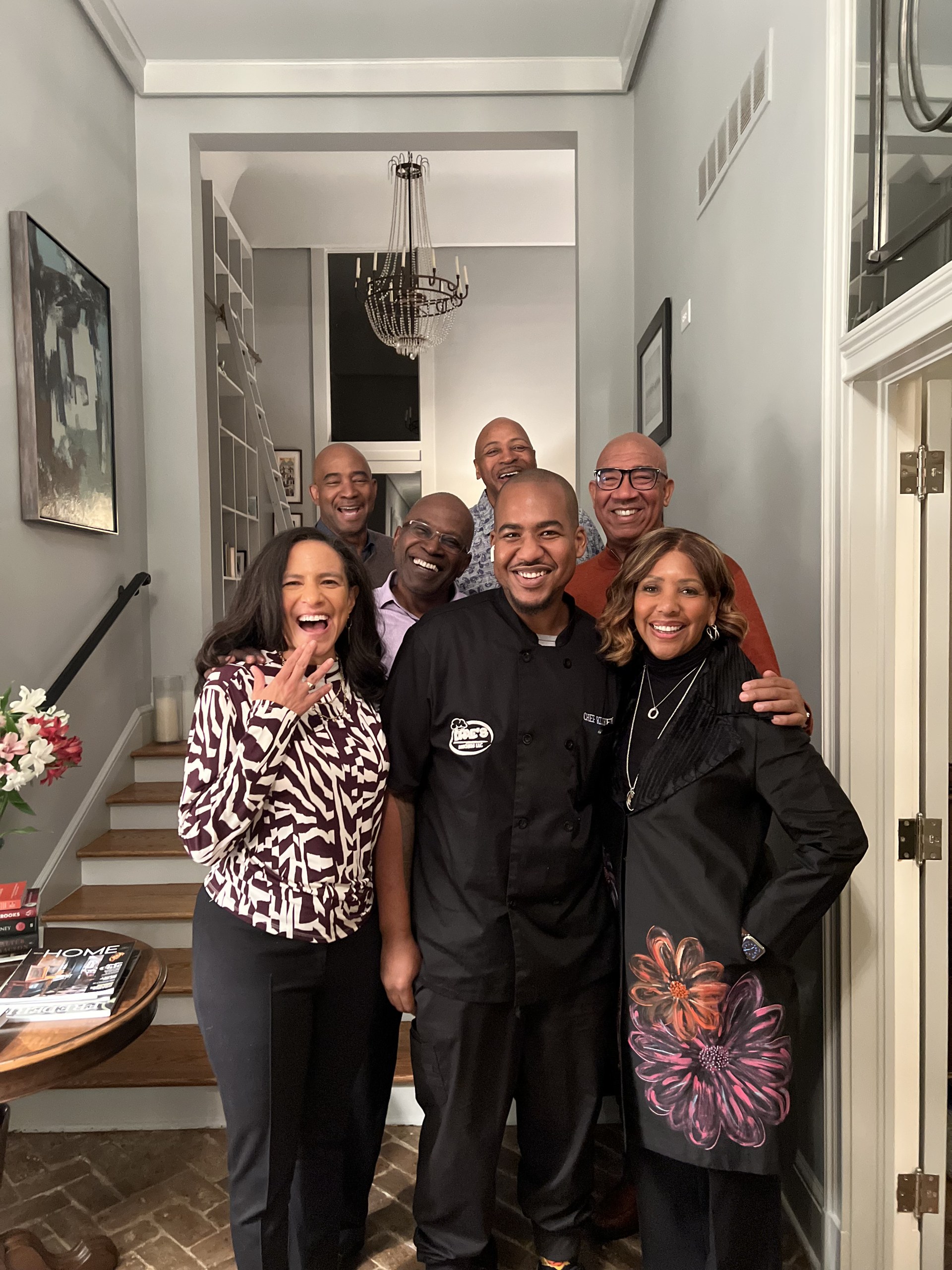 Chef Deandrae in a black chef coat smiling with a group of guests inside a home.