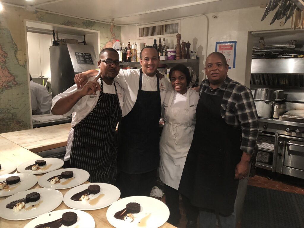Four people stand together in a working kitchen beside white plates with individual chocolate desserts and sauce.