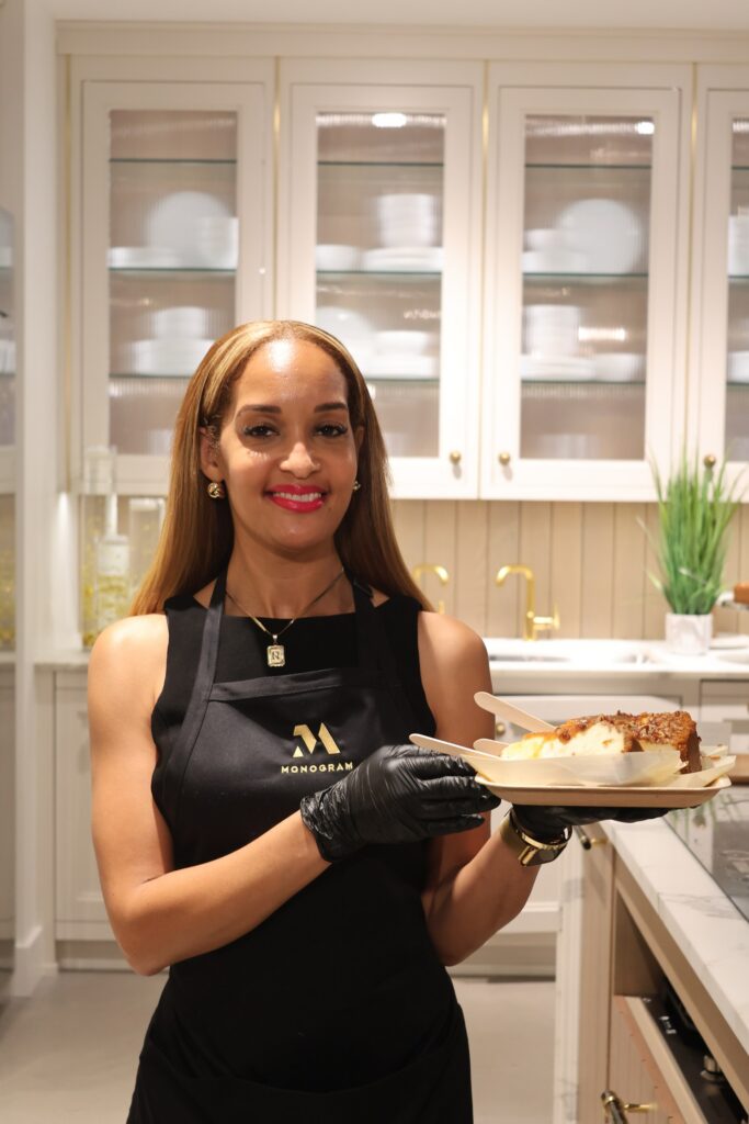A smiling woman in a black apron and gloves holds a tray with a slice of cheesecake in a bright kitchen with white cabinets.