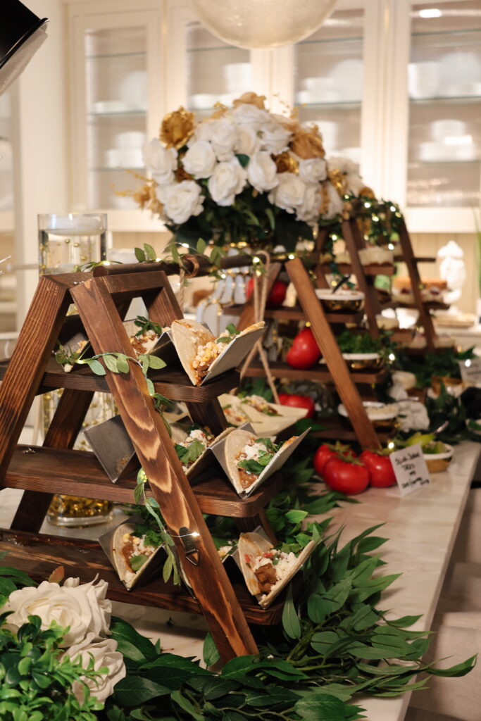 Individual tacos displayed on wooden tiered stands among white and blush flowers, leafy greenery and tomatoes.