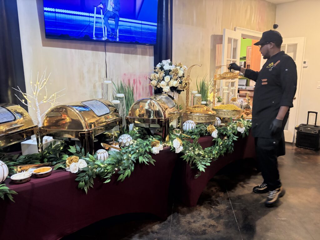 Chef Deandrae working beside a catered buffet with gold serving dishes, floral arrangements and a burgundy tablecloth.