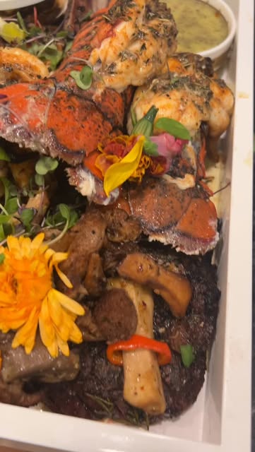 A catering platter of lobster tails and sliced meat garnished with edible flowers, herbs and vegetables.