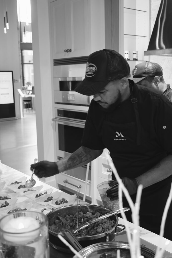 Black-and-white photograph of Chef Deandrae in a cap and apron, spooning food onto individual plates in a kitchen.