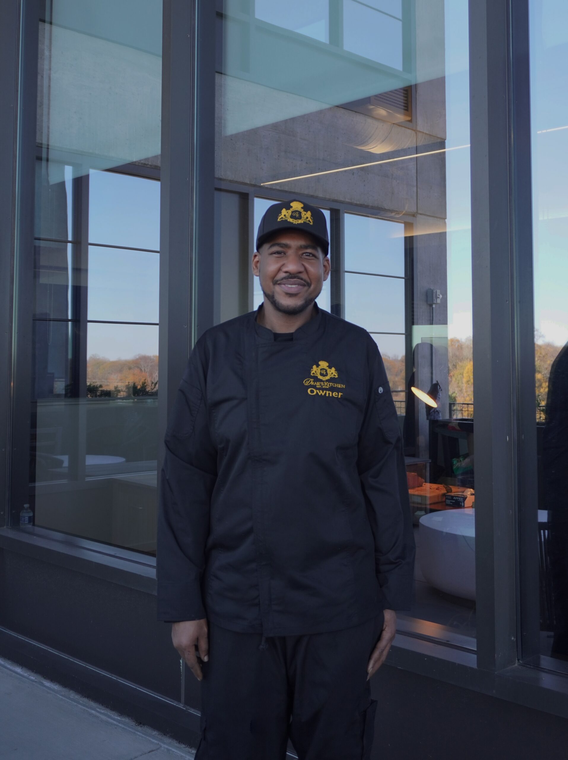 Chef Deandrae Kitchen in a black branded chef coat and cap, standing outside a glass-fronted building.