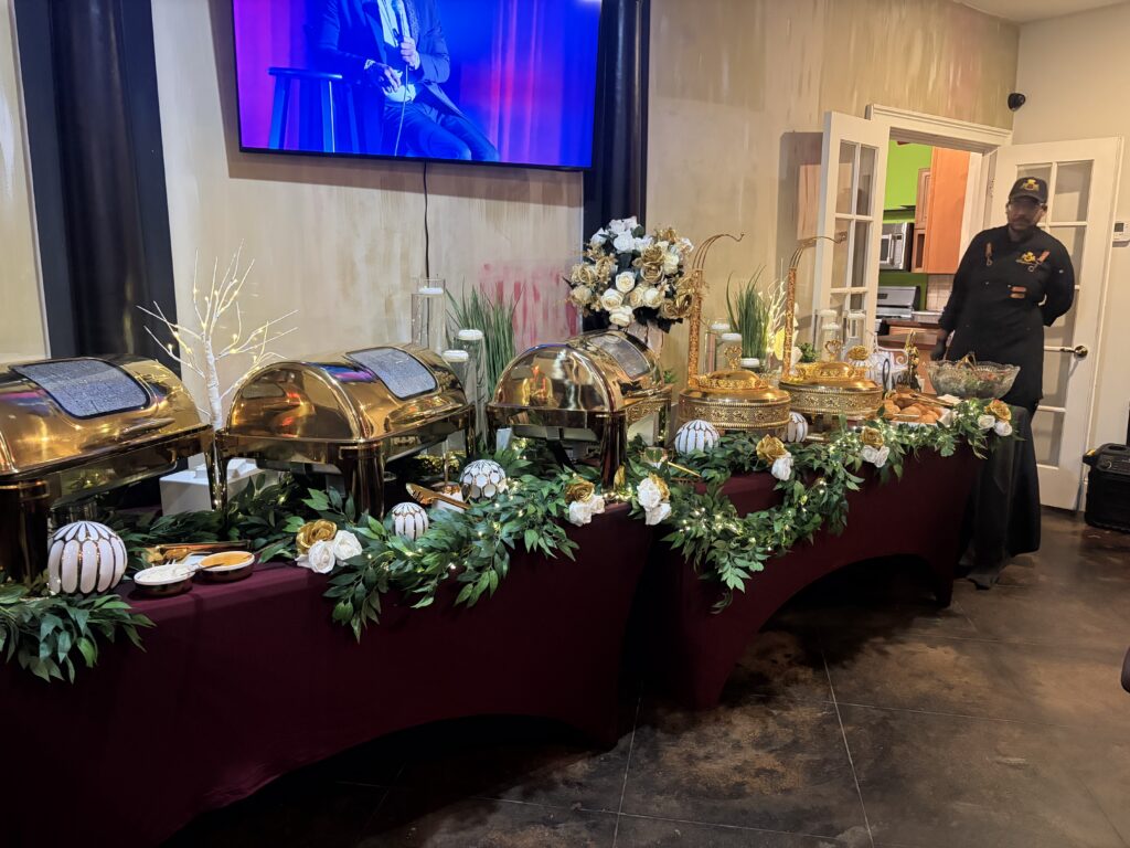 Chef Deandrae beside an event buffet with gold chafing dishes, burgundy tablecloths, white flowers and greenery.