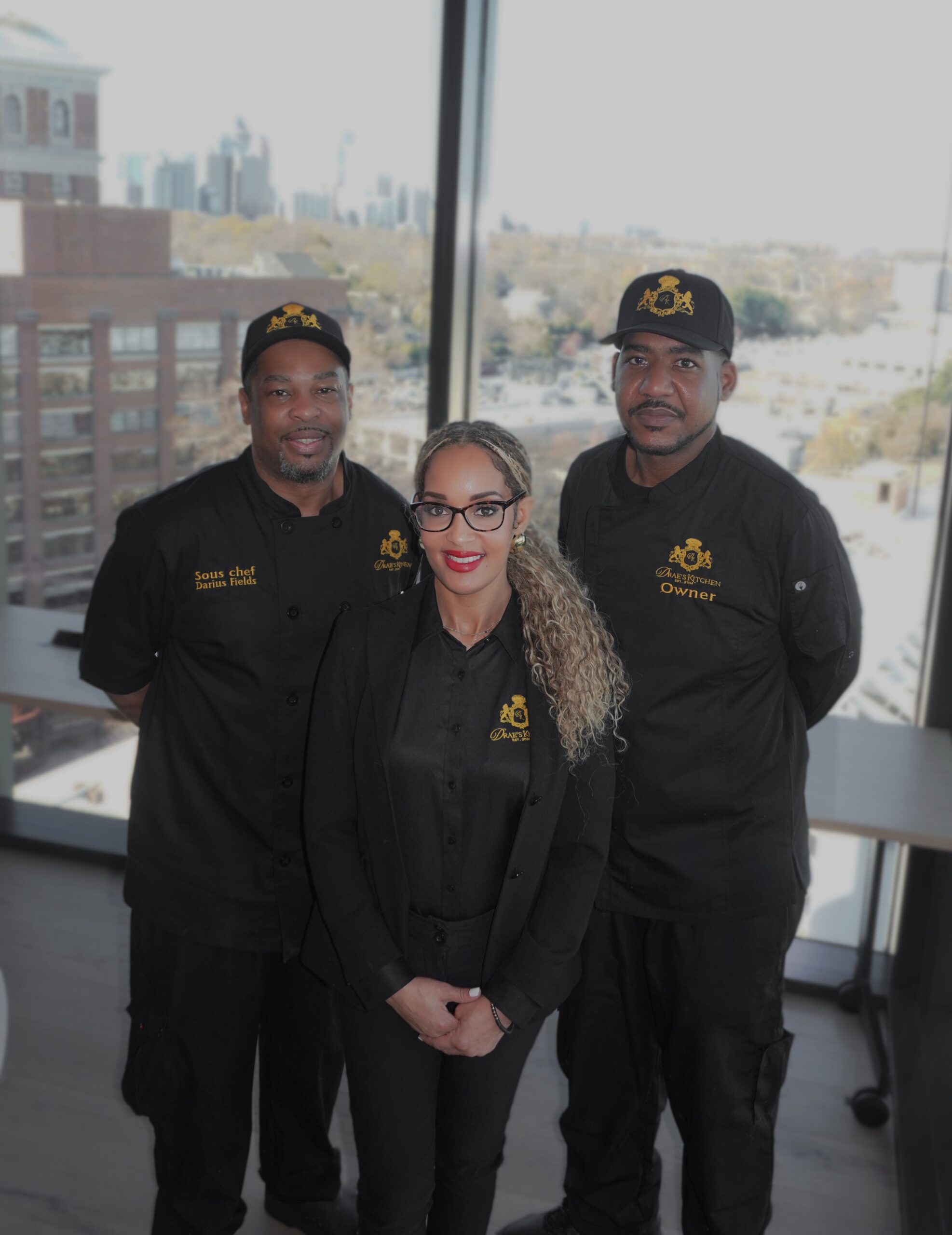 Three members of the Drae’s Kitchen team in matching black chef coats, standing together beside large windows.