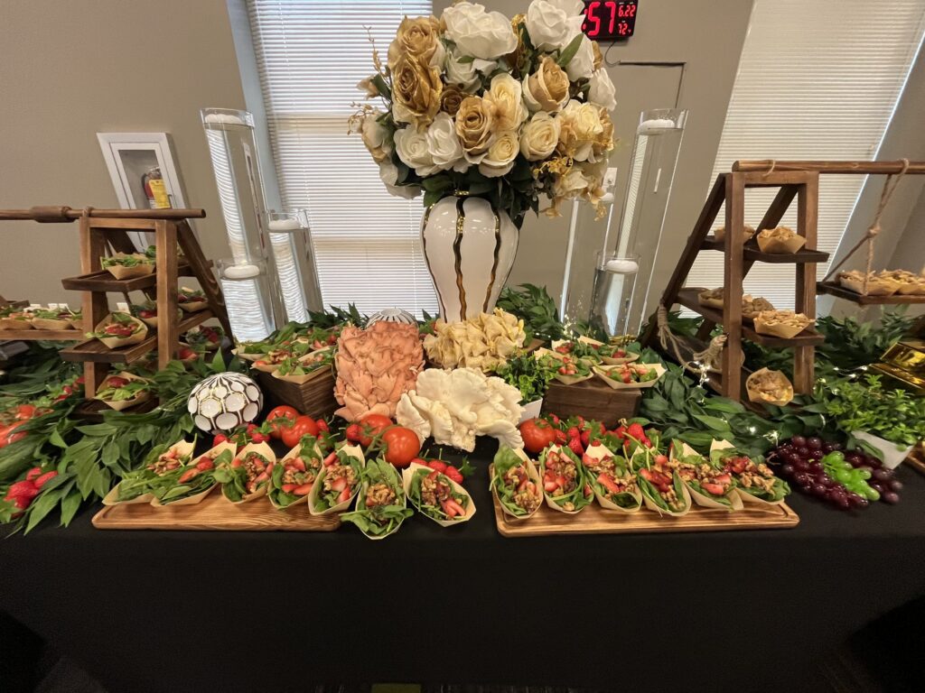 Tacos, a shrimp display, vegetables and fruit arranged beneath white-and-gold flowers, with wooden serving stands and greenery.